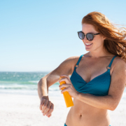 Happy young woman with red hair wearing sunglasses applying sun block cream on arm. Cheerful mature woman sunbathing while applying sunscreen lotion on body. Smiling lady in blue bikini with freckles on skin before sunbathing at beach with copy space.