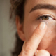 Close up of young woman wearing contact lens with copy space. Young woman holding transparent contact lens on index finger while looking at camera. Close up eye of healthy beautiful girl about to wear contact-lens.